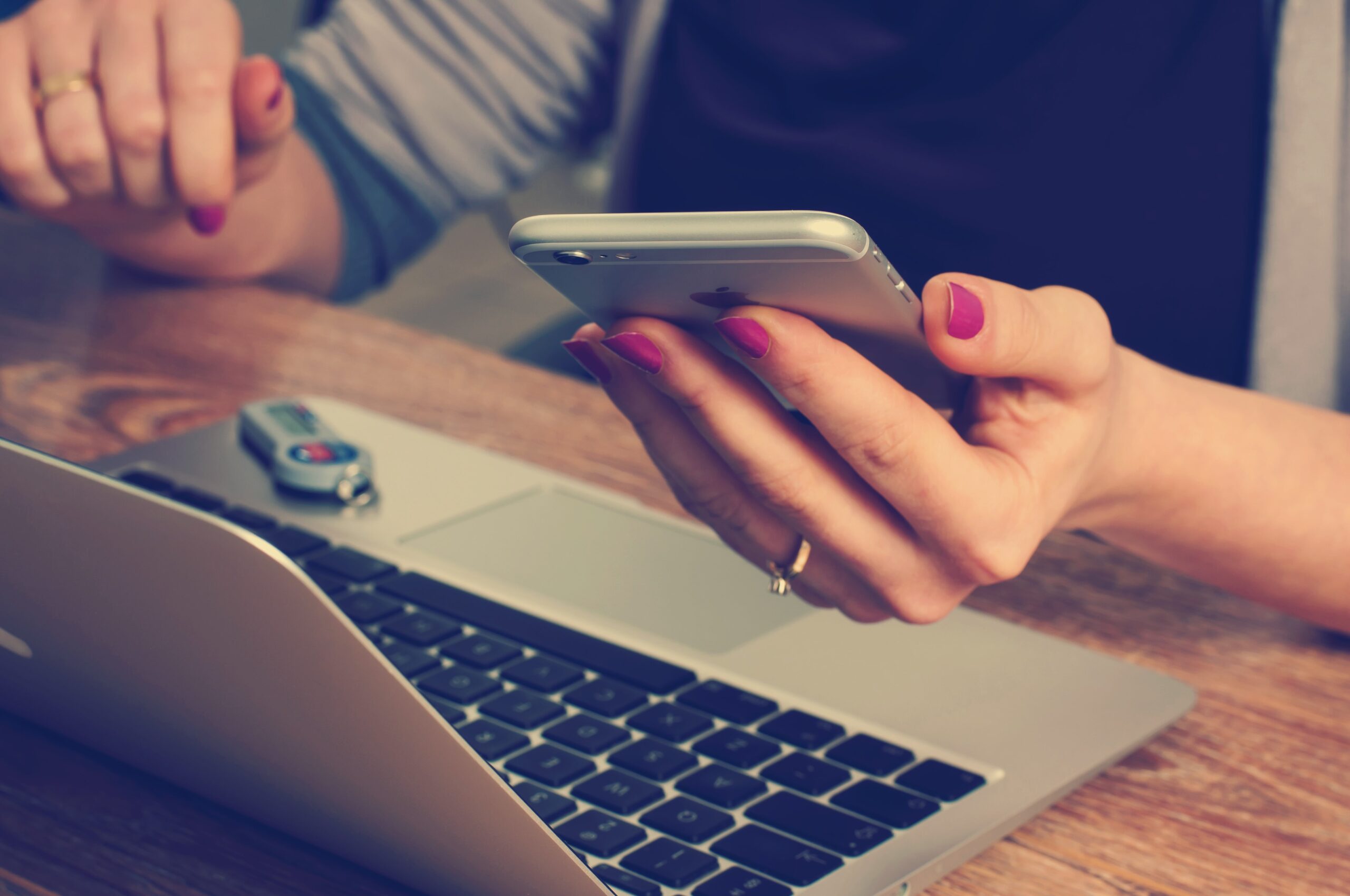 There is a woman holding an iPhone while sitting at a table in front of a laptop. She is wearing a grey sweater and black t shirt. Her fingernails are painted magenta, and she is wearing rings. This moment exemplifies the moment in the buying journey when consumers are seeking additional information about products from these brands, their differentiation in the market and perhaps are comparison shopping to understand which ones will provide good value and quality for their needs.