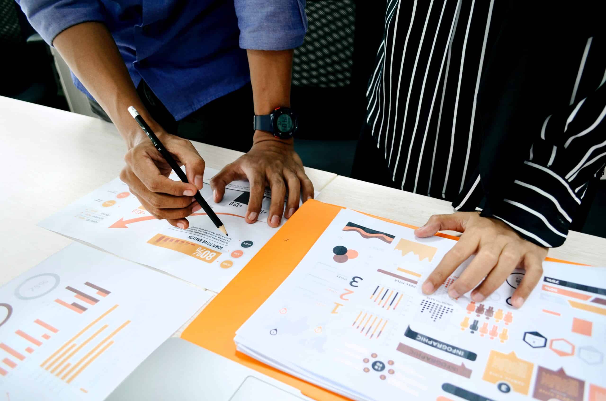 Two people standing in front of a table with several creative designs and marketing analytics dashboards in front of them.  They are collaborating to find the ideal solution for a client challenge.