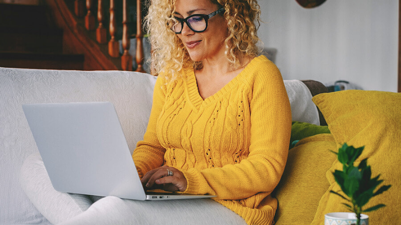 A woman wearing a yellow sweater and white leggings is sitting on a coach in her living room, using a laptop.