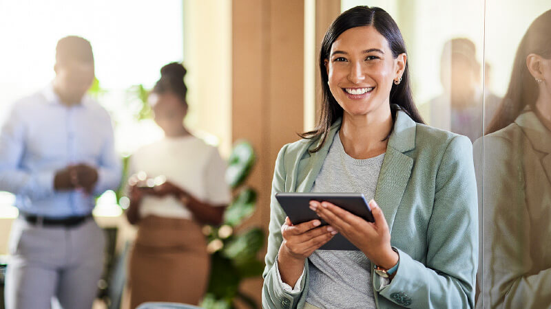 Professional woman standing in an office hallway against a glass wall with two coworkers standing in the distance.  She’s wearing a suit and holding an iPad.