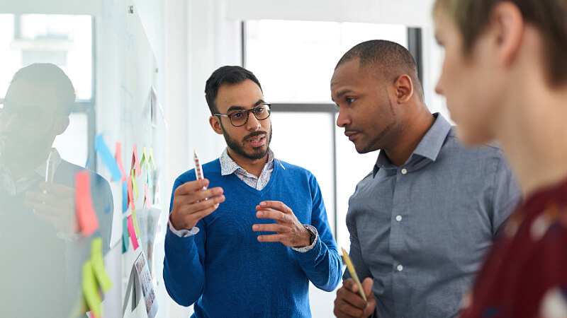 A team of professionals working in an office, standing in front of a glass board covered with notes and post-it notes. They are collaborating on the development and program implementation process.