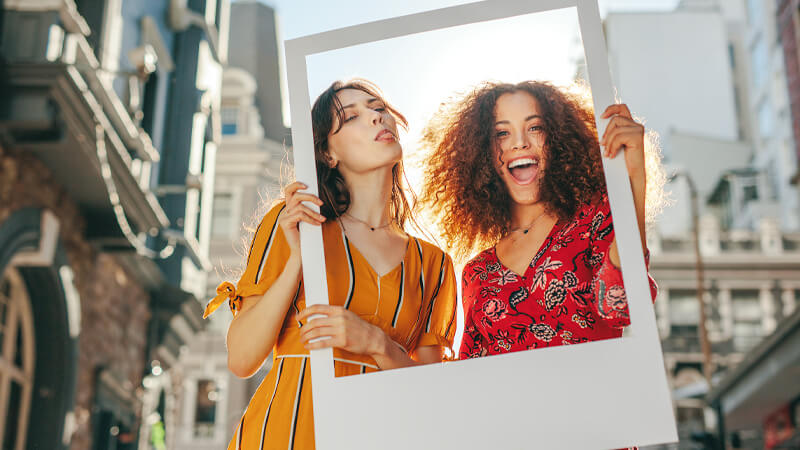 Two women posing outside for a picture while holding a cut out frame that resembles one from social media platforms.