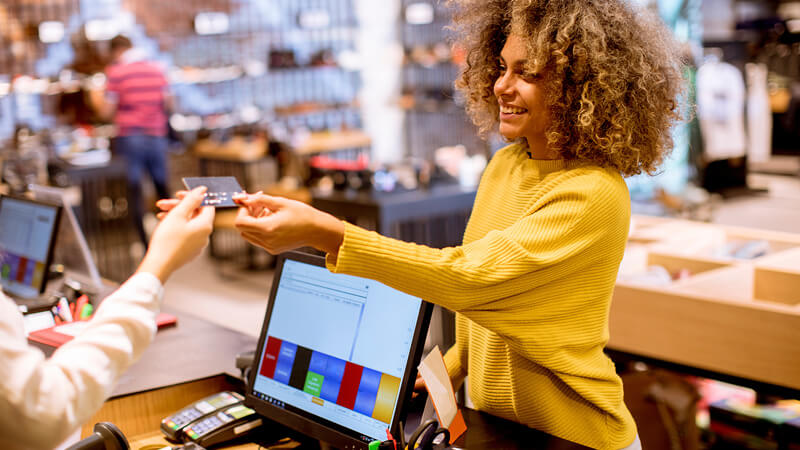 A woman wearing a yellow shirt handing her credit card to a store employee to purchase an item from a store.