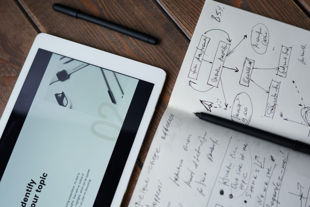 An open notebook that has writing on the pages and a pen placed on top with a tablet alongside it