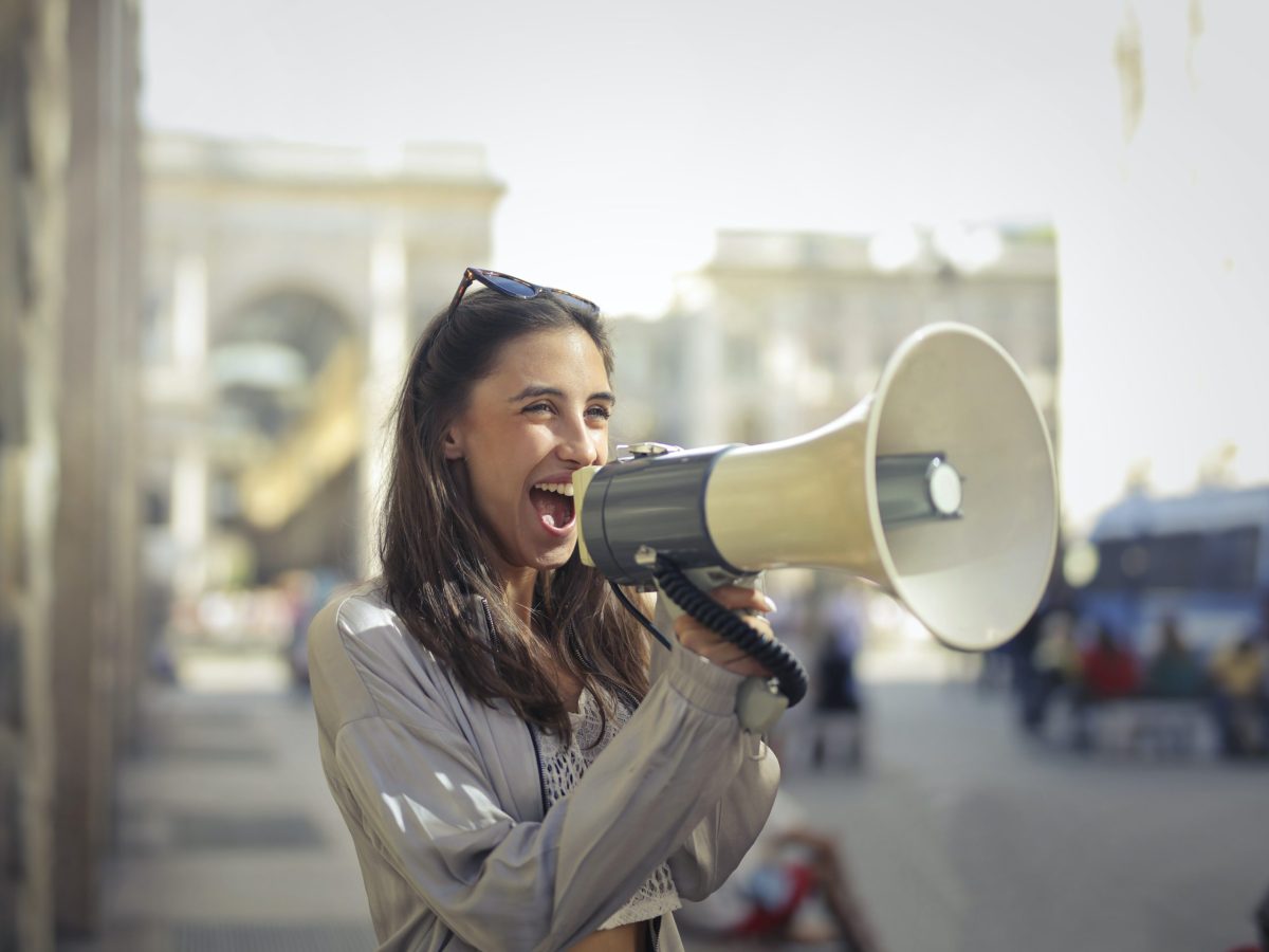 Woman standing on a busy, noisy and overly crowded street. She is holding a megaphone to amplify her voice so she can be heard by those that matter most.