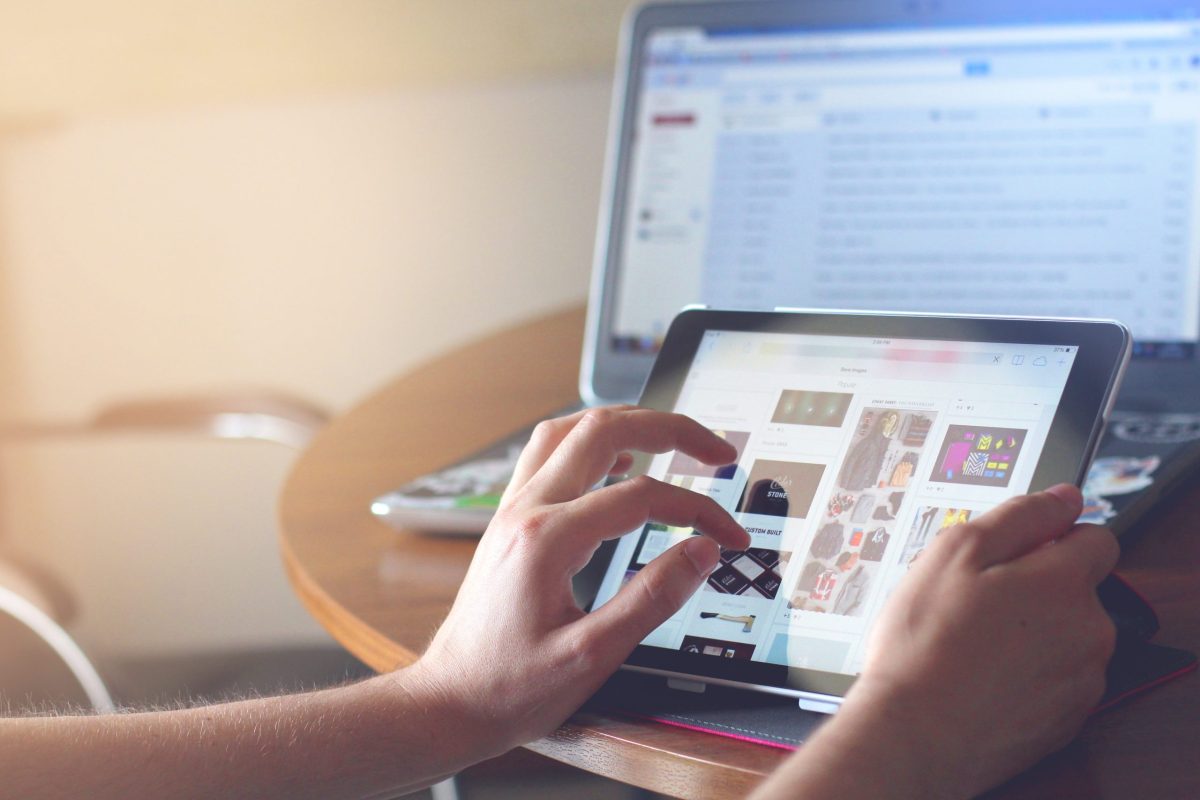 A hand scrolling on a tablet with a laptop sitting in the background