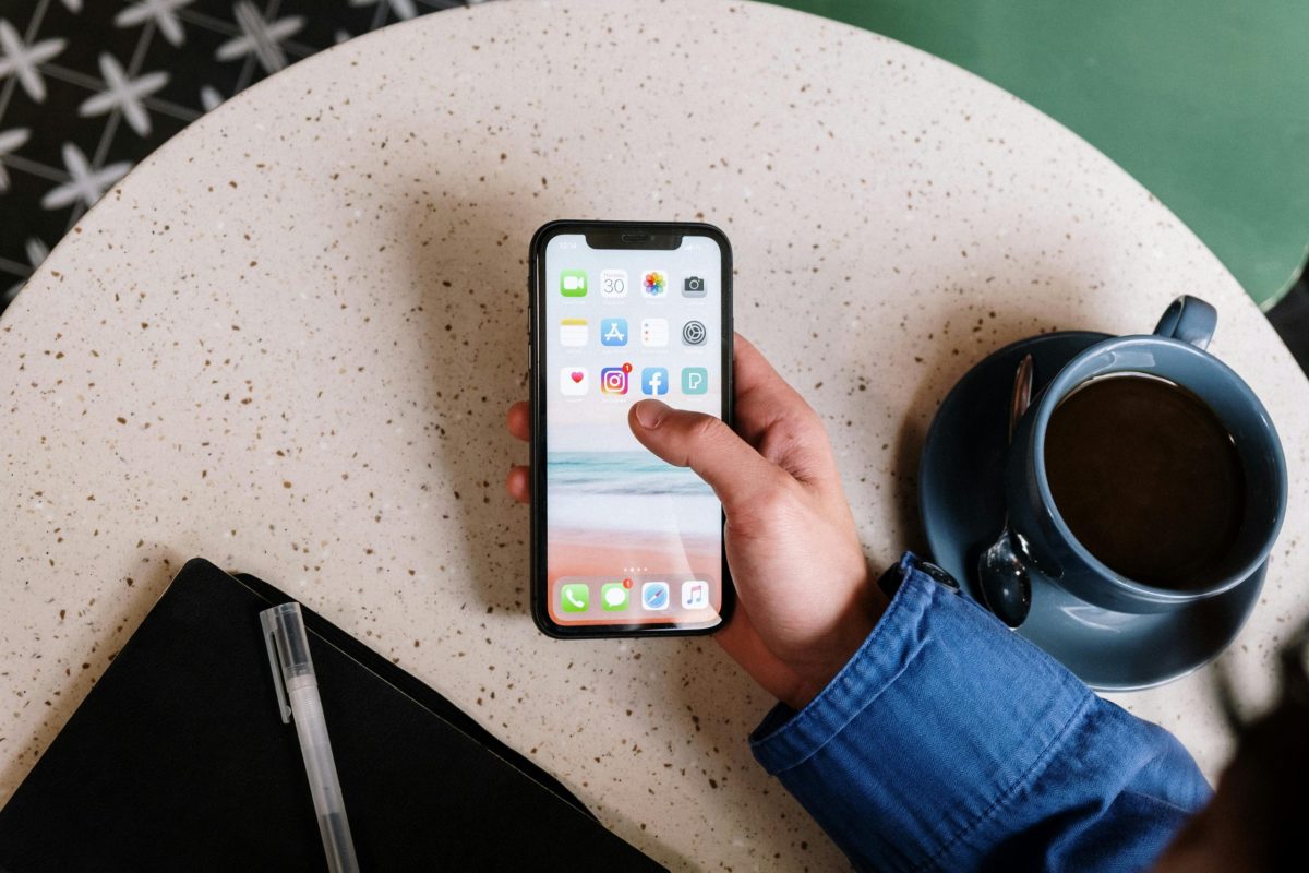 A person holding a smartphone at a cafe table with a coffee cup, a notebook, and a pen